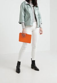 Světle zelená lesklá bunda, pruhované tričko, bílé úzké džíny, černé kotníkové boty a oranžová kabelka. Moderní, elegantní design s odvážným barevným kontrastem.