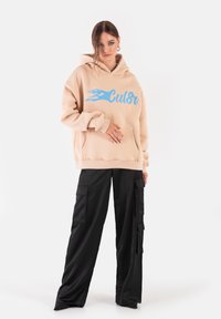 Beżowy hoodie z niebieskim wzorem płomienia i kieszenią z przodu, w połączeniu z czarnymi spodniami cargo z bocznymi kieszeniami i luźnym krojem.