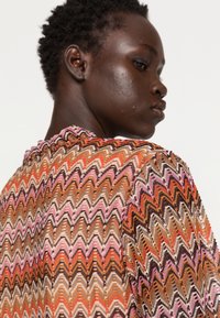 Top tricotado com um padrão em zig-zag em cores quentes: laranja, rosa, castanho e creme. Apresenta uma superfície texturizada e leve babado na gola.