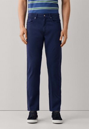 Hombre vestido con pantalones azul marino y zapatillas negras con suelas blancas, de pie frente a un fondo neutro.
