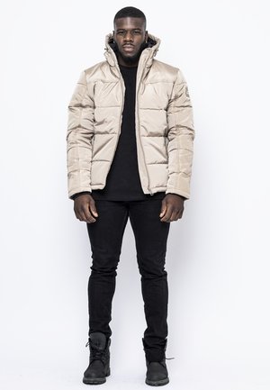 Chaqueta acolchada beige con cierre frontal y capucha, con un diseño acolchado. Llevada sobre una camiseta negra y combinada con jeans negros y botas.