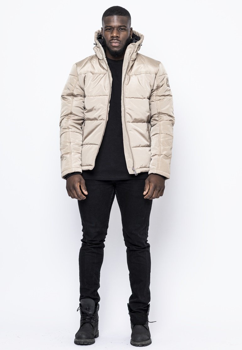 Giacca puffer beige con zip e cappuccio, caratterizzata da un design trapuntato. Indossata sopra una maglietta nera e abbinata a jeans neri e stivali.