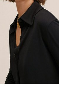 Chemise noire à col avec boutons, présentant une texture lisse et un design ajusté. Le col est pointu, accentuant l'aspect élégant.