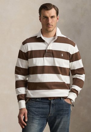 Mand iført en brun og hvid horisontalt striped rugbytrøje med hvid krave og blå jeans, stående med den ene hånd i lommen.