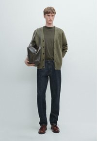 Junger Mann in olivgrünem Cardigan und T-Shirt, dunklen Jeans und braunen Schuhen, der eine große dunkle Leder-Umhängetasche vor einem schlichten Hintergrund hält.