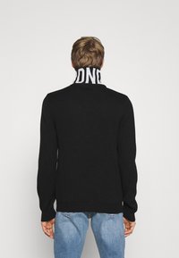 Černý pletený svetr s vysokým límcem, který má vzor bílého loga, žebrované manžety a spodní lem; střední hmotnost textilie.