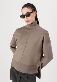 Brauner Rollkragenpullover mit lockerem Schnitt, geripptem Saum und seitlichen Schlitzen, kombiniert mit Schildkrötensonnenbrille. Glatte Textur, lässiges Design.