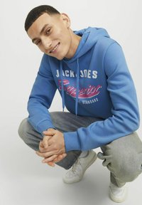 Sudadera azul con capucha y texto gráfico en blanco y rosa, combinada con jeans gris claro y zapatillas blancas. Atuendo casual, en pose de cuclillas.