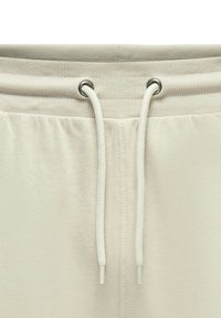 Pantalones de chándal de color beige claro con una cinturilla acanalada, que presenta ojales plateados y cordones blancos. Tejido suave con un ligero brillo.