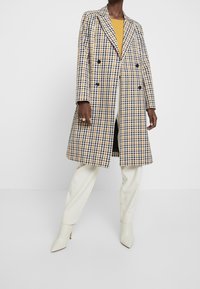 Dvoudílný kostkovaný kabát v námořnické modré, žluté a bílé. Obsahuje velké klopové límce a strukturovaný střih, spojený s krémovými kalhotami a podpatky.
