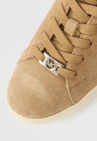 Beżowy zamszowy sneaker z okrągłymi sznurowadłami, srebrnym akcentem metalowym i teksturowaną cholewką. Gumowa podeszwa z delikatnym wzorem bieżnika.