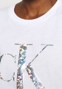Camiseta de algodón blanca con un gran logotipo de papel de aluminio iridiscente impreso en plata. El escote es de estilo clásico y tiene una textura suave.