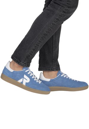 Personne portant un jean noir et des baskets en daim bleu avec des lacets blancs et un logo "R" blanc, debout avec un pied légèrement levé.