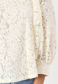 Blusa in pizzo color crema, con motivi floreali, maniche lunghe a palloncino e polsini elasticizzati, abbinata a jeans in denim. Tessuto testurizzato e leggero.