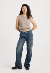 Top en tricot sans manches couleur khaki avec des boutons sur le devant, associé à un jean en denim indigo à coupe large et des bottes noires brillantes, le tout sur un fond uni.