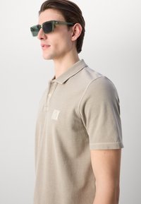 Beige polo skjorte med kort krage og tre knapper, med teksturert stoff og en liten logo på venstre bryst. Grønn solbriller.