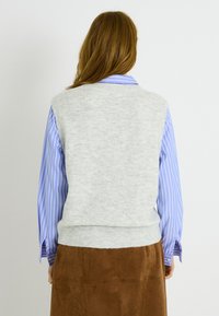 Chaleco de punto gris claro con cuello redondo, llevado sobre una camisa azul a rayas y una falda de ante marrón, visto desde atrás.