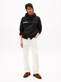 Felpa nera con strisce orizzontali bianche e rosse, indossata sopra una camicia a quadri rossi e neri, abbinata a pantaloni bianchi e scarpe slip-on marroni.