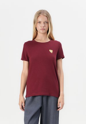 Lyshåret kvinde iført en burgundy t-shirt med et lille gyldent hjerte på brystet og løse grå bukser, stående mod en hvid baggrund.