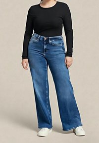Personne portant un haut noir à manches longues, un jean bleu taille haute et jambes larges, ainsi que des chaussures blanches, debout devant un fond clair uni.