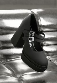 Μαύρο ψηλοτάκουνο παπούτσι με πλατφόρμα από λείο υλικό, με δύο διακοσμητικά λουράκια με ασημένιες αγκράφες, τοποθετημένο πάνω σε αντανάκλαση.