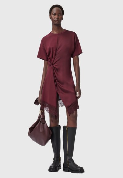 Robe courte bordeaux à manches courtes avec un détail noué à la taille et un ourlet en dentelle. Associée à des bottes noires à mi-mollet et un sac à épaule marron.