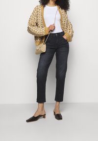 Cardigan testurizzato giallo senape con motivo geometrico bianco, abbinato a jeans scuri e mule nere con tacco, e una piccola borsa beige.