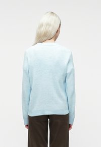 Cilvēks ar garām blondām matiem, ģērbies gaiši zila džemperī un tumšos biksēs, stāv ar muguru pret vienkāršu baltu fonu.