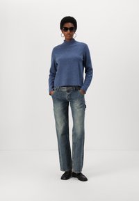 Blue, high-neck sweater paired with faded straight-leg denim jeans. Model wears black sunglasses and shoes, hands in pockets.