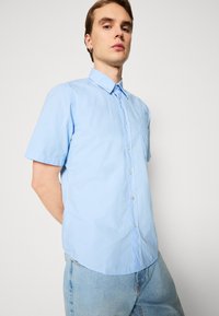 Camisa de botones de manga corta azul claro con cuello clásico, que presenta un bolsillo de parche y un ligero brillo, combinada con jeans de mezclilla.