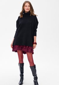 Zwarte oversized coltrui over een gelaagde bordeauxrode jurk met kantaccenten. Zwarte kniehoge laarzen maken de look compleet.