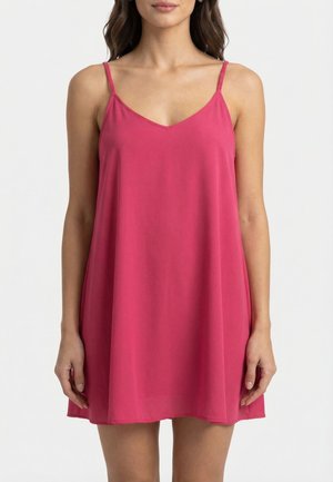 Femme portant une robe sans manches, ample et rose vif avec de fines bretelles, sur un fond clair uni.