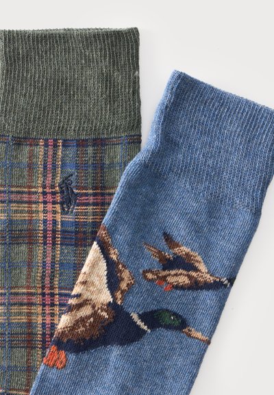 Deux chaussettes tricotées, l’une avec un motif à carreaux verts et marron, l’autre bleue avec des dessins de canards en vol, toutes deux montrées depuis la zone du poignet.