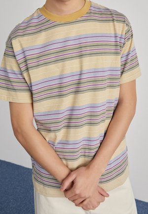 Person iført en kortærmet gul, lilla og sort stribet t-shirt med en sennepsfarvet krave, hænderne foldet foran.