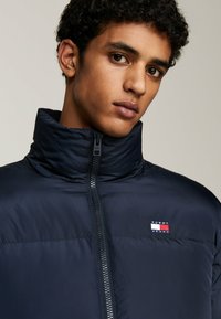 Giacca imbottita blu scuro con colletto alto, chiusura con zip e texture impressa. Presenta una piccola toppa con il logo "Tommy Jeans" sul petto.