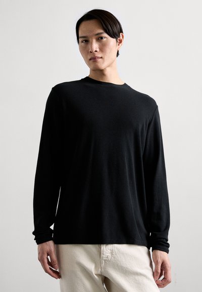 Tricou negru cu mânecă lungă, realizat dintr-un material moale și texturat, cu guler rotund. Purtați-l cu pantaloni de o nuanță deschisă, potriviți pe încheieturi.