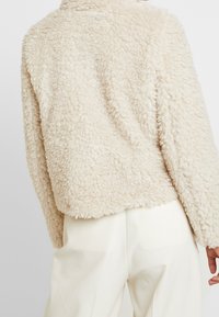 Krämfärgad, plyschig, faux fur-jacka med en mjuk textur och avslappnad passform. Har långa ärmar och en rundad nederkant.