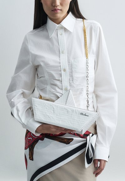 Kobieta nosząca białą bluzkę z marszczeniami i drukowany szalik, trzymająca białą torebkę w kształcie papierowej łódki z złotym i srebrnym łańcuchem.