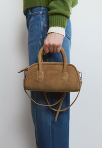 Mano sosteniendo un pequeño bolso de ante marrón con asas y una correa desmontable, contra un fondo de jeans azules y un suéter verde.