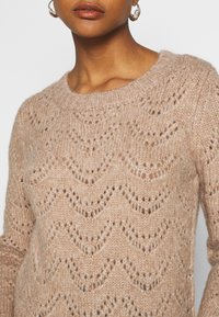 Suéter de punto en beige claro, con un patrón de olas y un cuello redondo. Hecho de un hilo suave y texturizado con un corte holgado.