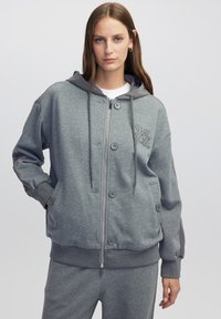 Sudadera gris con cremallera fabricada en un tejido suave, que cuenta con botones, capucha y puños acanalados. Incluye un sutil bordado en la parte delantera.
