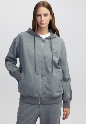 Sudadera gris con cremallera fabricada en un tejido suave, que cuenta con botones, capucha y puños acanalados. Incluye un sutil bordado en la parte delantera.