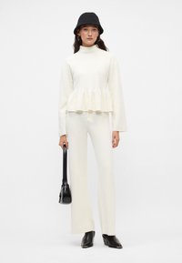 Maglione bianco di lana, con texture e design a peplum, abbinato a pantaloni dritti coordinati. La modella tiene una borsa nera e indossa un berretto di maglia nero.