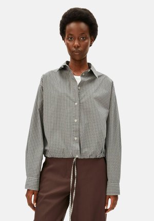 Femme aux cheveux courts et bouclés portant une chemise à carreaux grise à boutons avec une taille resserrée par un cordon et un pantalon marron sur un fond clair uni.