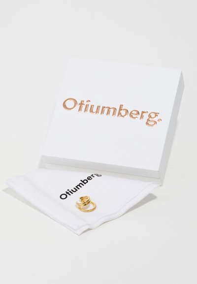 Guldring på et hvidt stof med "Otiumberg" branding, placeret ved siden af en hvid æske med roséguld prægede bogstaver.