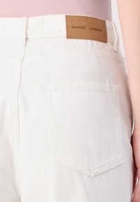 Jeans en denim blanc avec une étiquette en cuir portant "SAMSØE SAMSØE" et une poche arrière avec des coutures visibles sur le côté gauche.