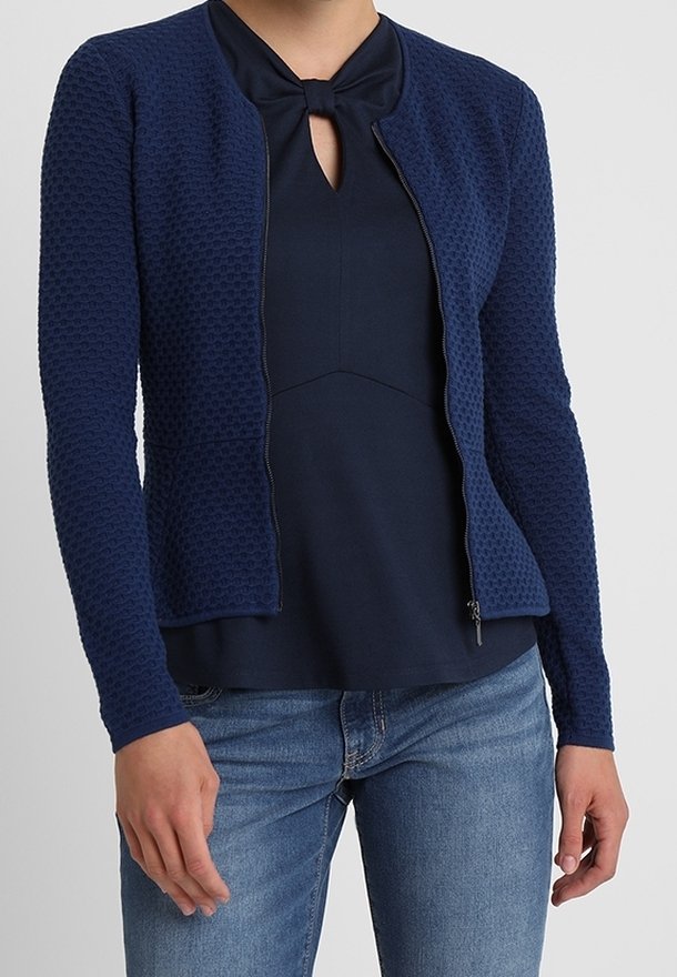 Femme portant un cardigan zippé texturé bleu marine par-dessus un haut bleu marine avec un détail nœud, assorti à un jean bleu, sur un fond uni.