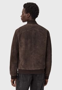 Brązowa zamszowa kurtka bombowca z prążkowanymi mankietami i dół, z tylnym szwem yoke i gładką teksturą, połączona z ciemnymi dżinsami.