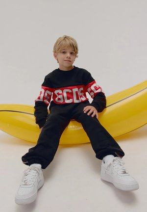 Felpa nera con striscia rossa e lettere bianche, pantaloni sportivi neri, sneakers bianche, seduto su un grande oggetto di scena giallo a forma di banana.