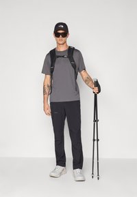 Abbigliamento da escursionismo con una maglietta grigia, pantaloni leggeri neri, zaino nero, scarpe grigie e un cappellino nero con occhiali da sole. Bastoncino da trekking in mano.
